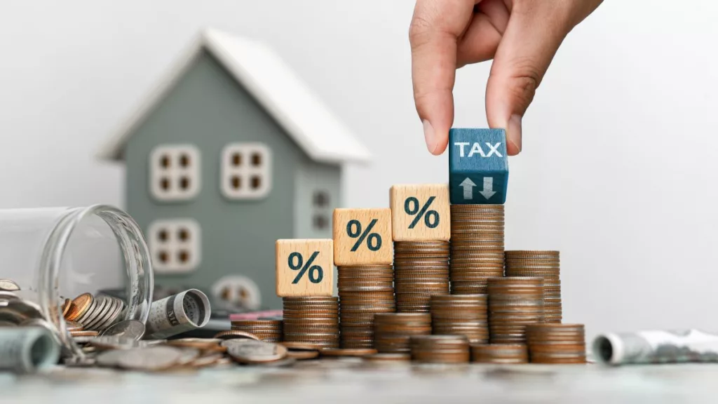 A hand places a "TAX" block on growing stacks of coins beside a house model, symbolizing property tax increases and deferrals under the California Property Tax Postponement Program.